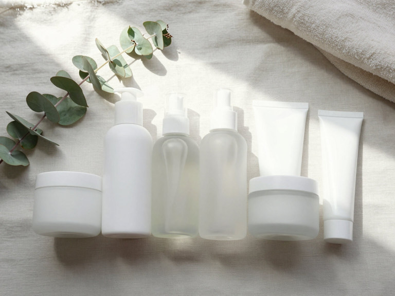 An overhead flat-lay of unlabelled skincare bottles and tubes on linen with eucalyptus.