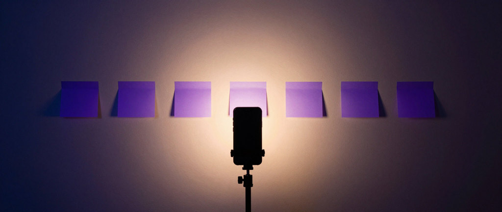 Seven violet notes in a row on a wall at dusk, a phone on a tripod lit from behind.