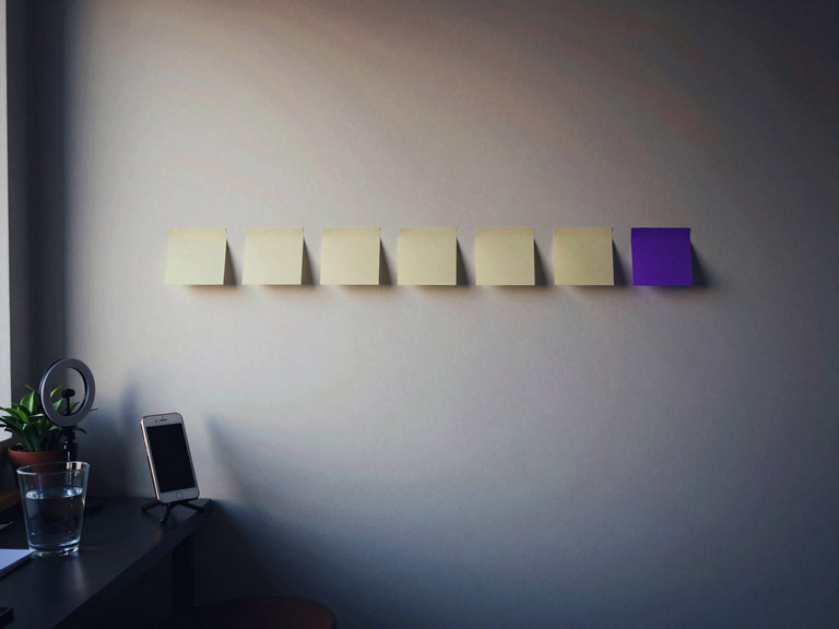 A creator's corner in the morning: seven sticky notes in a row on the wall, the last one violet, and a phone on a small tripod below.