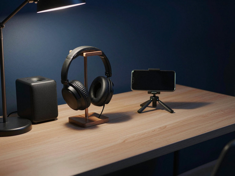 Headphones on a stand and a small speaker on a desk in the evening, a phone on a mini tripod filming.